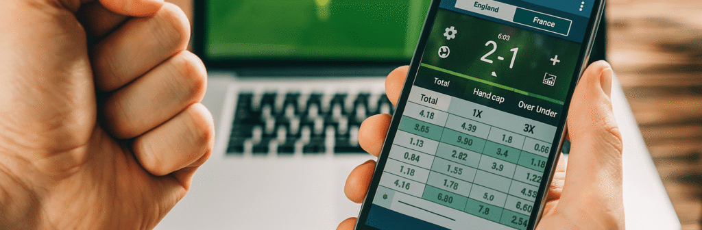 paris-sportif-bookmaker-etranger-mobile Application de bookmaker étranger affichant un score en direct entre l'Angleterre et la France sur un smartphone, devant un écran d’ordinateur
